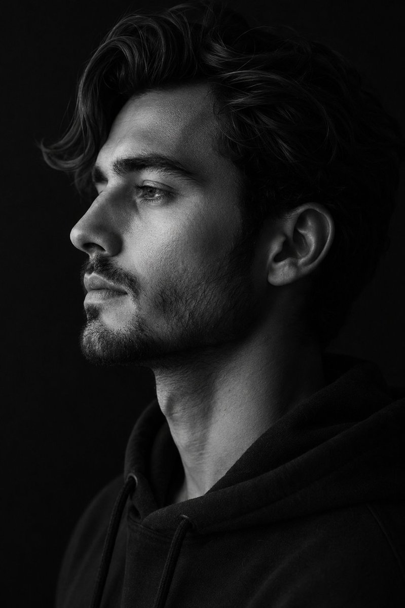 A single figure turned fully to the left against a deep black backdrop, lit so the jawline, brow and lips read as crisp sculpted edges. Soft light lifts the…