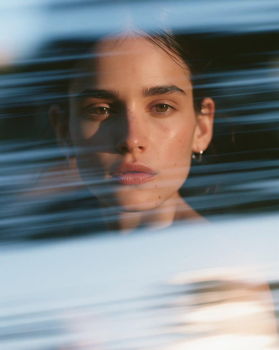 A sharp, calm close-up of a woman with a low bun and silver hoops, while translucent blue strips streak across the foreground in soft motion. The face stays…