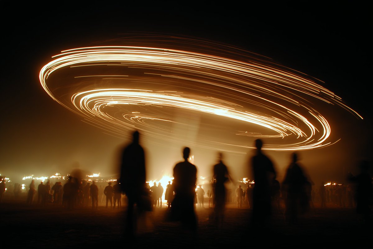 A spinning ferris wheel burns against a dark night sky while small figures watch from below. The fire, the wheel's silhouette and the crowd's stillness give…