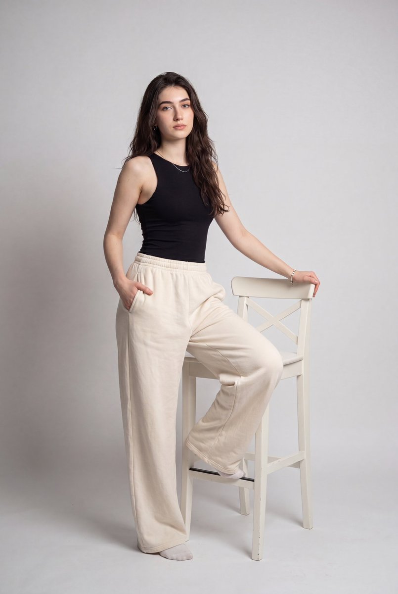 A full-length portrait of a woman with long dark hair in a fitted black tank and oversized cream wide-leg pants, one leg resting on a white bar chair. Soft…