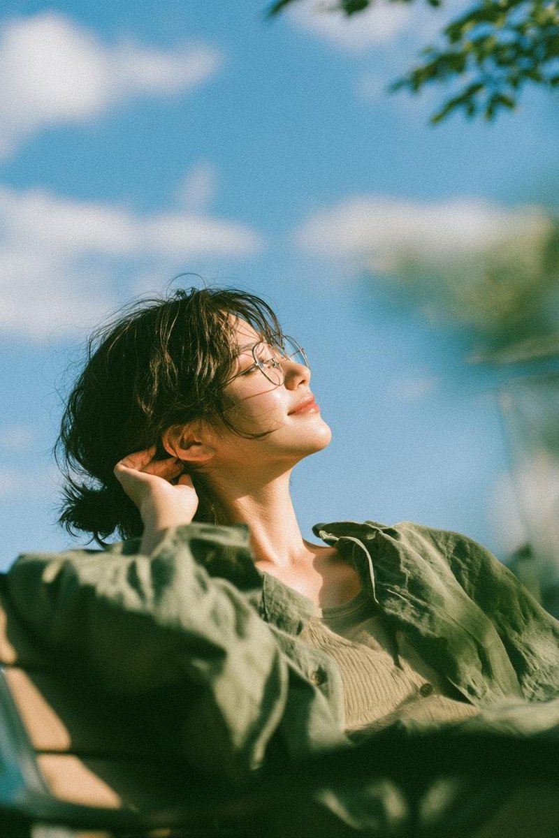 A young woman with a short black bob rests in a leafy garden as warm sunlight washes over her face. The soft olive linen, blue sky, and creamy blur give it a…