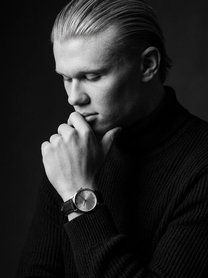 A tight, upright crop of a man in profile, eyes lowered and one hand raised to his face, with a watch catching light in the lower left. Deep shadow fills the…