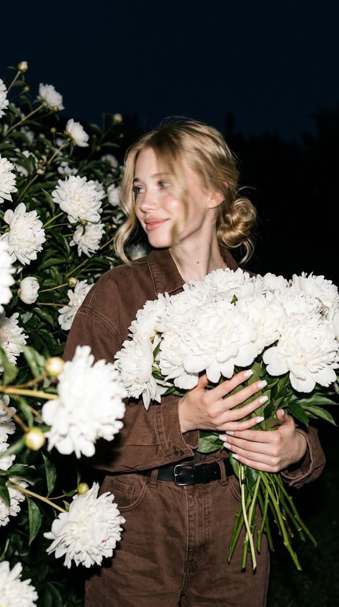 A young woman in a chocolate-brown denim jumpsuit stands among white peonies after dark, lit by a crisp on-camera flash against a deep navy sky. The result…