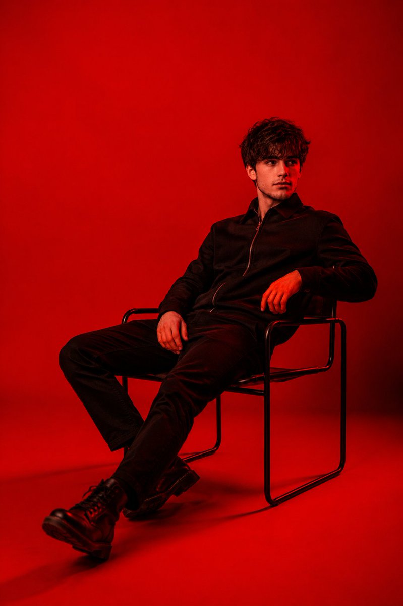 A man in an all-black jacket, pants and boots sits back on a thin metal chair against a seamless backdrop of saturated red. The high contrast and calm pose…