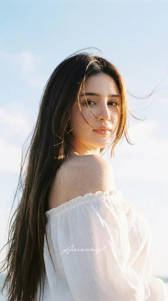 A soft, sun-washed portrait of a woman seen from behind, glancing over one shoulder. Loose strands lift in the breeze, backlight rims her hair and shoulder…