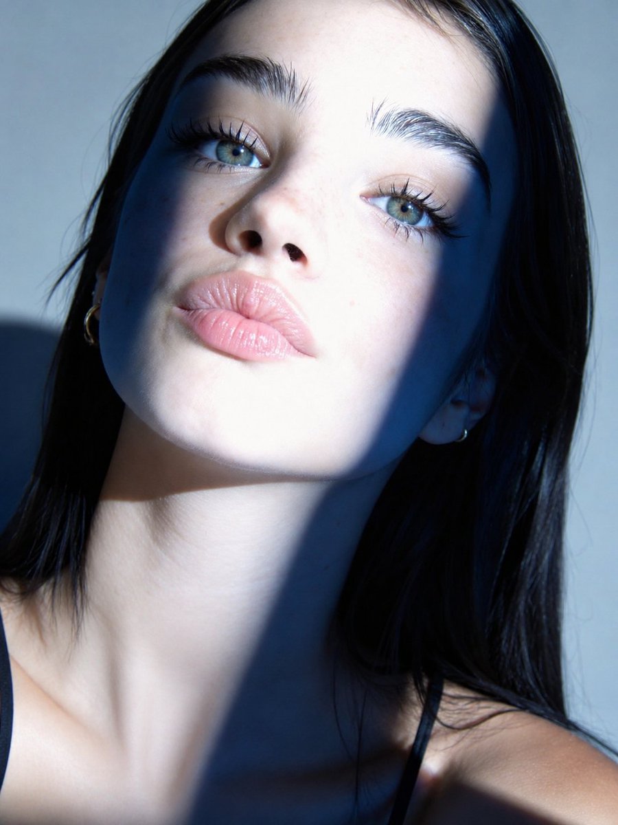 A tight, face-filling beauty shot with a diagonal window shadow splitting the frame into cool and golden halves. Freckles, glossy nude lips and blue-green eyes…